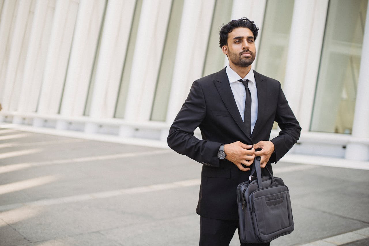 Business professional in a suit buttoning jacket while holding a briefcase outside a modern building.